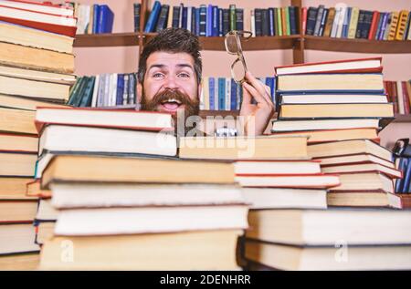 Homme sur le visage heureux entre des piles de livres dans la bibliothèque, des étagères sur fond. Professeur ou étudiant avec barbe porte des lunettes, assis à la table avec des livres, démoqué. Concept de recherche scientifique. Banque D'Images