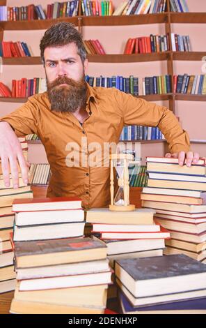Concept de bibliothécaire. Professeur, scientifique avec la barbe se tient à table avec des livres, défoqué. L'homme sur le visage réfléchi se tient entre des piles de livres, tout en étudiant dans la bibliothèque, les étagères sur le fond. Banque D'Images