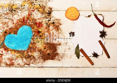 Concept de l'art alimentaire. Composition du condiment faisant la forme du coeur rempli de sel bleu. Morceau de papier avec bâtonnets de cannelle, étoiles anis et tranches d'orange sèches. Ensemble d'épices sur fond de bois beige. Banque D'Images