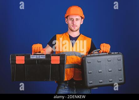 Homme dans un casque, casque de sécurité tient la boîte à outils et la valise avec des outils, fond bleu. Concept de la boîte à outils et de l'équipement. Travailleur, réparateur, réparateur, constructeur sur le visage réfléchi choisissant l'équipement pour le travail. Banque D'Images