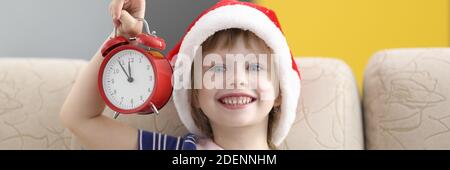 Une petite fille dans le chapeau du père noël est assise sur un canapé souriant et tenant un réveil. Banque D'Images