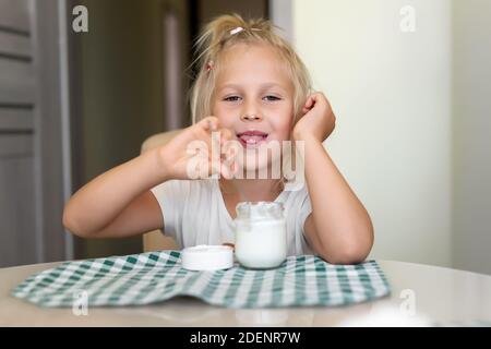 Mignon adorable caucasien petite fille blonde drôle manger du yogourt ou du fromage cottage de lait pour le déjeuner snack. Les enfants aiment manger assis à une table sur la cuisine Banque D'Images