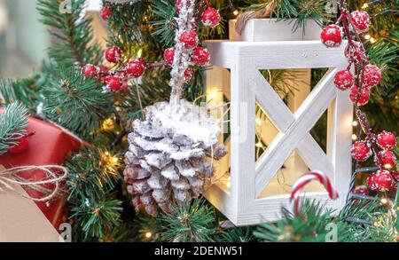 Lanterne en bois blanc avec guirlande, cône de pin enneigé et baies rouges sur un arbre de Noël. Banque D'Images