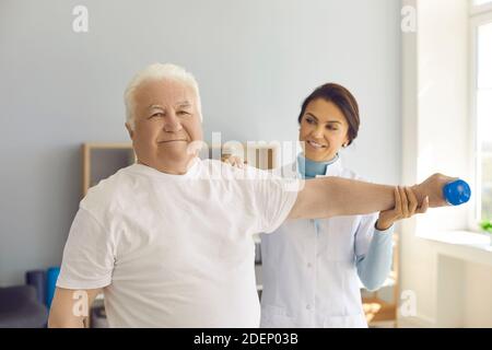 Sourire homme âgé patient faisant de l'exercice de physiothérapie avec haltère sous femme chiropraticiens contrôle Banque D'Images