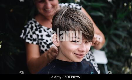 un jeune garçon obtient sa coupe de cheveux par une femme heureuse dans le jardin. Il est heureux et regarde la caméra. Banque D'Images