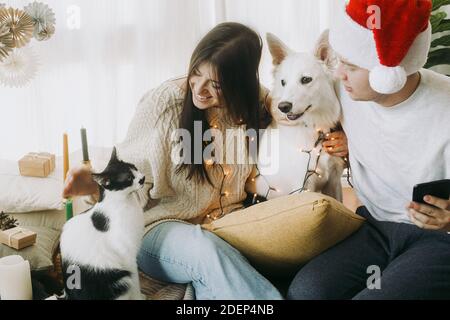 Joyeux jeune famille assis avec un chien mignon et chat dans une salle de boho décorée de fête, des moments doux. Joyeux Noël ! Couple élégant avec animaux de compagnie dans lumières et Banque D'Images