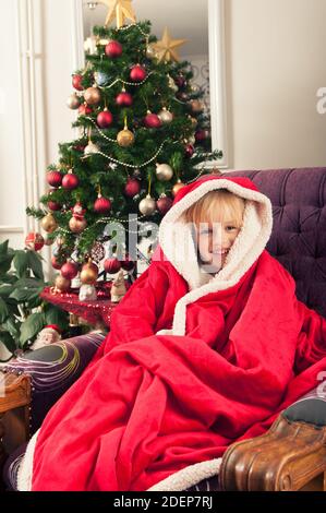 Belle petite fille avec chapeau de père noël enveloppé dans une couverture rouge, assis à côté de l'arbre de christmass et souriant Banque D'Images