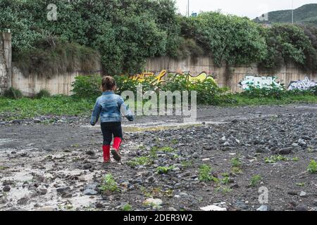 Fille sautant dans des flaques boueuses portant des bottes rouges, se remplissant de boue Banque D'Images