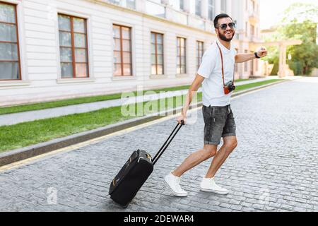 Touriste positif avec une barbe, avec une valise et un appareil photo, courant dans les rues, vers le repos Banque D'Images