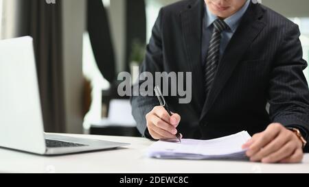 Un homme d'affaires confiant en costume noir écrit des notes ou un rapport financier dans un bureau moderne. Banque D'Images