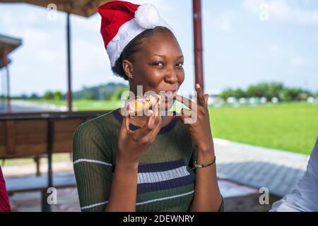 Un portrait en gros plan d'une femme noire attrayante dans un Chapeau de père Noël mangeant un délicieux donut et léchant son doigt Banque D'Images