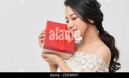 Bonne femme asiatique en robe tenant une boîte cadeau et regardant à l'appareil photo tout en bénéficiant d'un arrière-plan blanc Banque D'Images