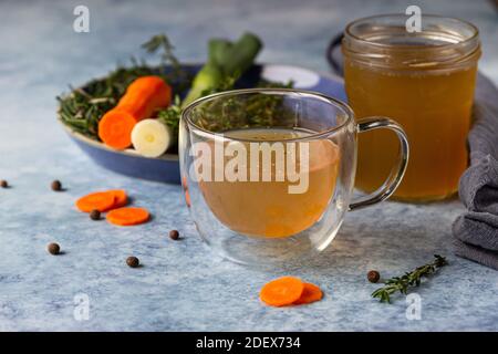 Bouillon d'os fait maison dans une tasse en verre et des légumes, fond en béton bleu. Source de collagène pour le corps. Mise au point sélective. Banque D'Images