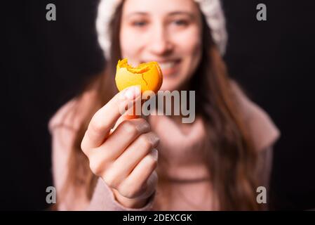 Le macaron jaune mordu est tenu par une jolie fille souriante sur fond noir. Banque D'Images