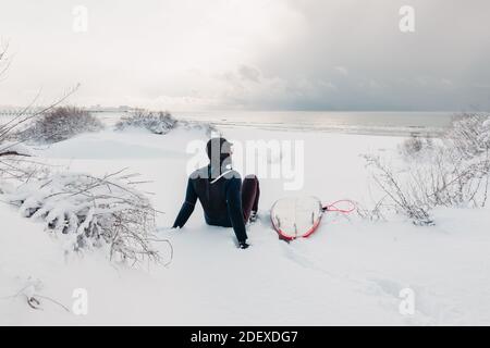 Hiver froid et surfeur assis sur la plage de neige avec planche de surf. Jour d'hiver avec surfeur en combinaison. Banque D'Images