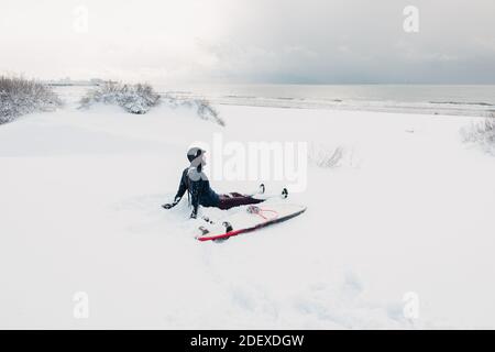 Hiver froid et surfeur assis sur la plage de neige avec planche de surf. Jour d'hiver avec surfeur en combinaison. Banque D'Images