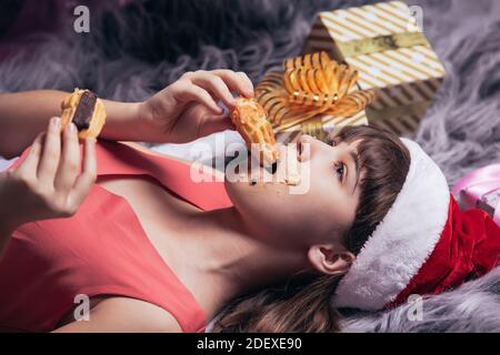 Une adolescente dans un chapeau de père Noël mange des gâteaux de Noël sur le tapis, dans les boîtes dorées de fond de cadeaux. Photo de haute qualité Banque D'Images