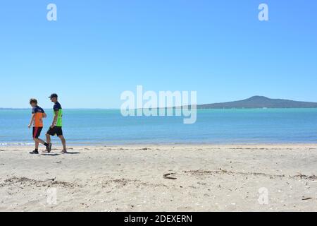 AUCKLAND, NOUVELLE-ZÉLANDE - 28 novembre 2020 : vue de deux jeunes hommes marchant sur la plage de St Heliers avec l'île Rangitoto en arrière-plan Banque D'Images