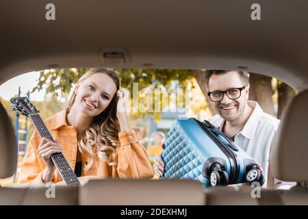 Couple souriant avec guitare acoustique et valise regardant la caméra près du coffre de la voiture sur un premier plan flou Banque D'Images