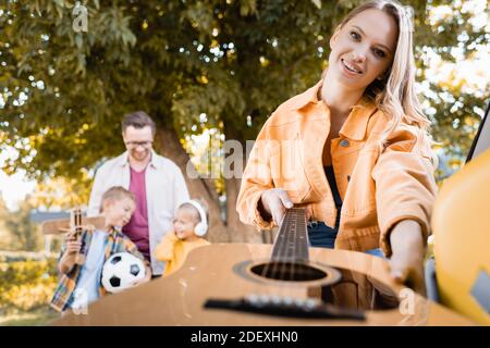 Femme souriante prenant de la guitare acoustique du coffre de voiture près de son mari et les enfants sur un arrière-plan flou en extérieur Banque D'Images