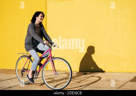 une jeune adolescente heureuse sourit alors qu'elle roule son vélo rose rétro le long d'un mur jaune coloré, concept de mode de vie actif et de mobilité durable, c Banque D'Images