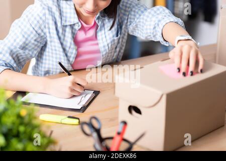 Vue rognée de l'écriture de volontaire féminin sur la planchette à pince tout en la tenant boîte en carton au bureau sur un premier plan flou Banque D'Images