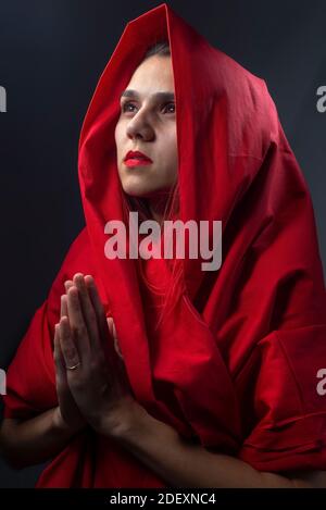 Portrait religieux dramatique fille en raies rouges avec les mains pliées. Studio photo sur fond gris belle sorcière asiatique avec cape rouge priant Banque D'Images