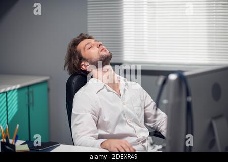 Un médecin de sexe masculin assis à son bureau dans une clinique médicale, se penchait sur la tête de la chaise et se reposant les yeux. Banque D'Images