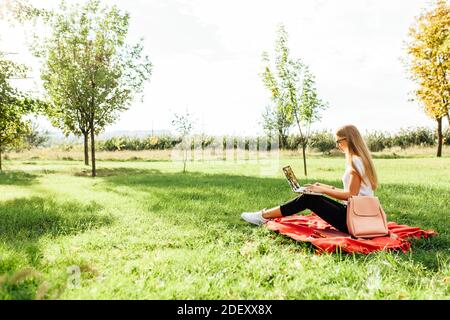 L'image d'une belle jeune fille étudiante avec des lunettes, qui travaille sur un ordinateur portable, assis sur une couverture rouge dans le parc faisant ses devoirs Banque D'Images