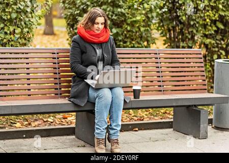 Jeune femme travaillant sur un ordinateur portable assis sur un banc de parc avec du café. Utilisation d'ordinateurs et d'achats en ligne. Formation à distance, formation en ligne Banque D'Images