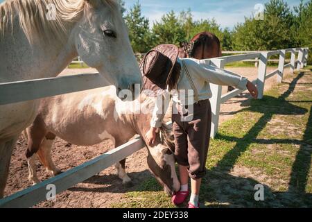 Bonne vie de ferme. Petite fille dans un stand de tenue vintage près du paddock avec un beau cheval blanc et petting un poney. La fille est en parie Banque D'Images