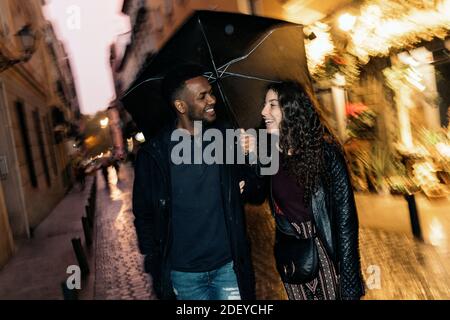 Photo de stock du moment heureux de couple interracial s'amuser sous la pluie dans la rue. Banque D'Images