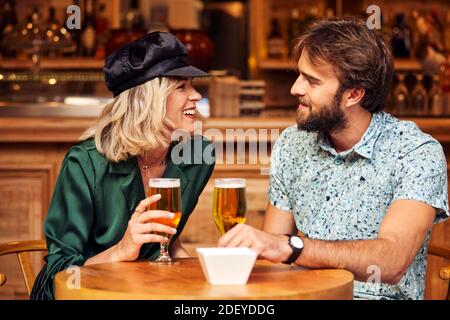 Photo de stock d'un couple dans leurs années 30 ayant un verre dans un bar. Ils sourient et rient. Ils portent un tissu décontracté. Banque D'Images