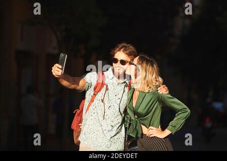 Photo d'un couple dans leur 30s marchant dans une rue. Ils prennent un selfie. Ils portent un tissu décontracté. Banque D'Images