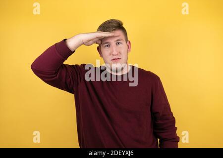 Jeune homme blond portant un chandail rouge décontracté sur fond jaune très heureux et souriant regardant loin avec la main sur la tête. Concept de recherche. Banque D'Images