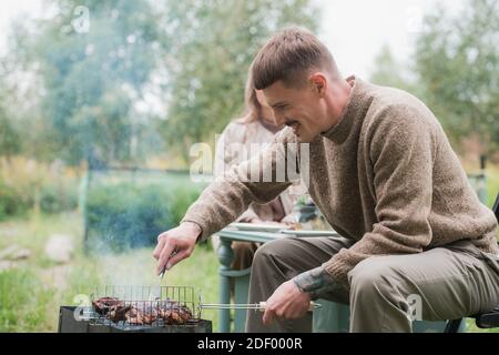 Le mari, le chef de famille, prépare des steaks et un barbecue pour sa femme lors d'un dîner à l'extérieur. Sur la pelouse dans la cour d'une maison de campagne Banque D'Images