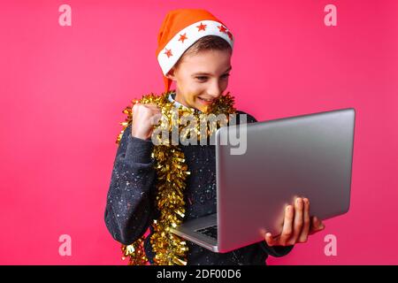Joyeux adolescent dans le chapeau de Santa et avec le tinsel sur son cou regardant l'ordinateur portable excité au sujet de la bonne nouvelle sur l'Internet, le gars heureux montre un geste de Banque D'Images