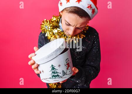 Un jeune garçon étonné dans le chapeau du Père Noël et avec une guirlande autour de son cou, ouvrant avec intérêt une boîte cadeau pour Noël, sur un fond rouge Banque D'Images