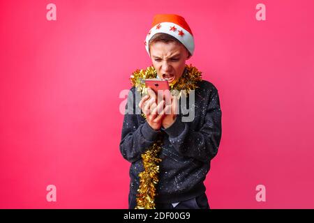 Portrait de l'adolescent en colère, dans le chapeau du Père Noël et avec la guirlande sur son cou, debout criant au mobile isolé sur fond rouge Banque D'Images