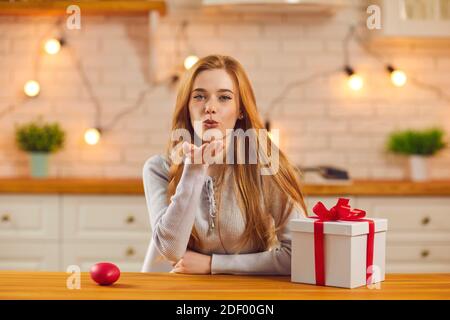 Jolie jeune femme assise à la table de cuisine avec un cadeau de Saint-Valentin et un baiser à l'appareil photo Banque D'Images