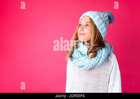 adorable adolescente en chandail blanc debout isolée sur fond rose portant un chapeau bleu chaud et une écharpe chaude. Concept, hiver, froid Banque D'Images