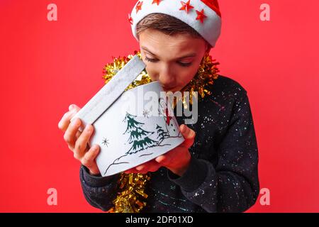 Un jeune garçon étonné dans le chapeau du Père Noël et avec une guirlande autour de son cou, ouvrant avec intérêt une boîte cadeau pour Noël, sur un fond rouge Banque D'Images