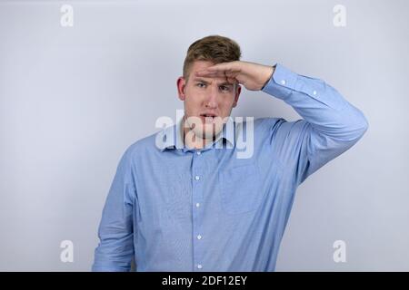 Jeune homme d'affaires portant une chemise décontractée sur fond blanc très heureux et souriant regardant loin avec la main sur la tête. Concept de recherche. Banque D'Images