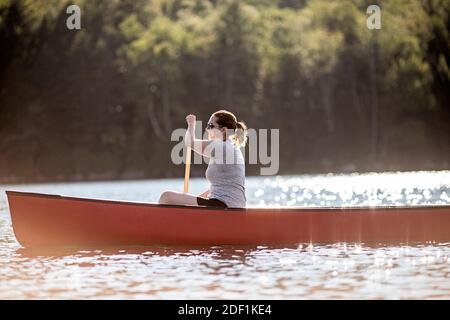 Femme pagaie rouge canoë à travers le lac sous le soleil dans le Maine bois Banque D'Images