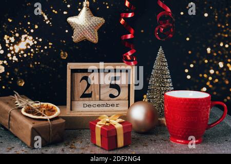 Calendrier en bois avec date du 25 décembre, décorations de Noël, tasse de thé rouge et boîtes-cadeaux sur fond noir avec des étincelles. Mise au point sélective Banque D'Images