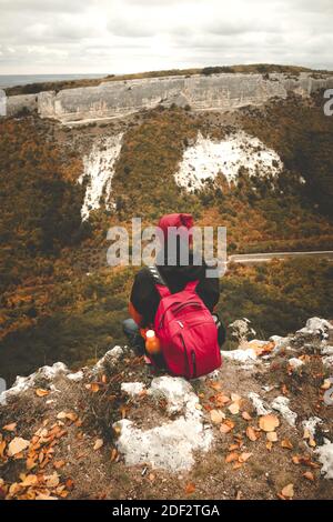 Voyageur assis sur le bord avec sac à dos rouge et bonnet, se détendre et profiter du paysage. Vue sur le paysage vertical de la forêt de montagne d'automne Banque D'Images