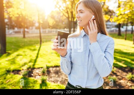 Une jeune fille, vêque officiellement, boit du café, écoute de la musique avec des écouteurs sans fil et marche dans le parc Banque D'Images