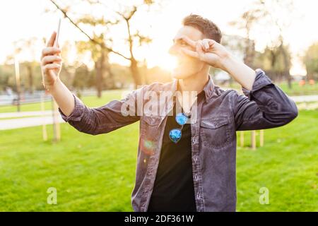 Portrait d'un homme en train de rire, portant des lunettes de soleil, debout dans un parc avec un smartphone. Il prend des photos de lui-même et montre le geste de paix sur c Banque D'Images