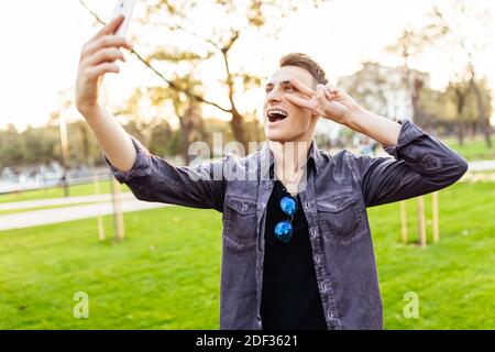 Portrait d'un homme en train de rire, portant des lunettes de soleil, debout dans un parc avec un smartphone. Il prend des photos de lui-même et montre le geste de paix sur c Banque D'Images