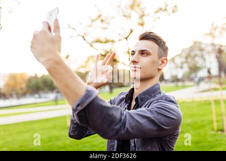 Portrait d'un homme heureux, en lunettes de soleil, debout dans le parc avec un smartphone. Il prend des photos de lui-même Banque D'Images